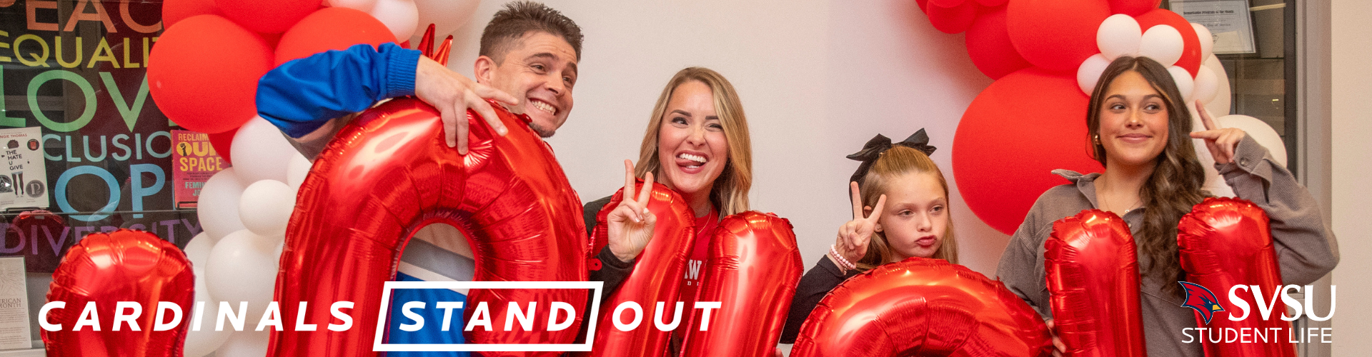A family standing with inflatable SVSU letters looking past the camera smiling and making peace signs with their hands.